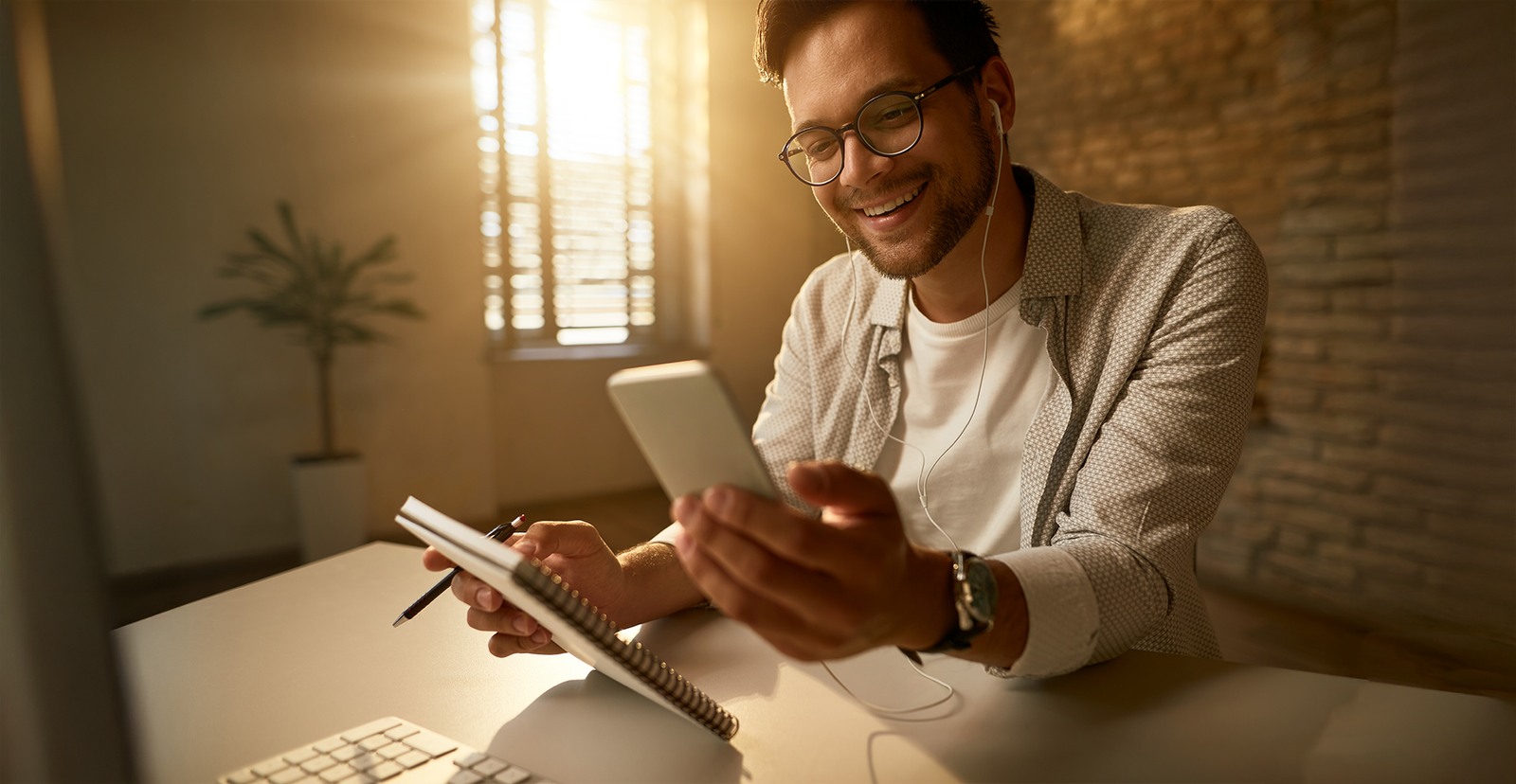 Un emprendedor sonriente con audífonos, revisando su teléfono móvil y tomando notas en una libreta, representando la eficiencia del trabajo con automatización de marketing.